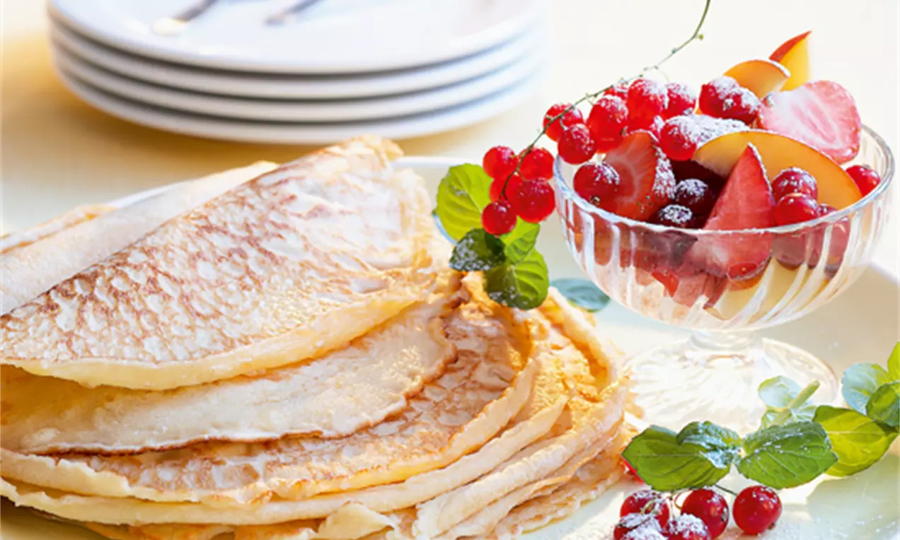 Crêpes fines et dorées empilées sur une assiette blanche, accompagnées de fruits rouges et jaunes frais dans une coupe transparente
